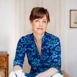 Mariana Leky sitzt im blau-gblümnten Oberteil vor einer hellen Wand.
