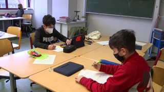 Zwei Schüler sitzen an einem Gruppentisch mit Mund-Nasen-Bedeckung.