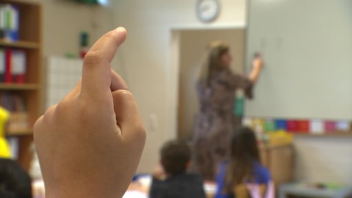 Ein Schulkind meldet sich im Unterricht, die Lehrerin ist an der Tafel. 