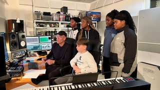 EIne Gruppe junger Schülerinnen sitzt mit einem Musikproduzenten im Tonstudio und blickt auf die Monitore