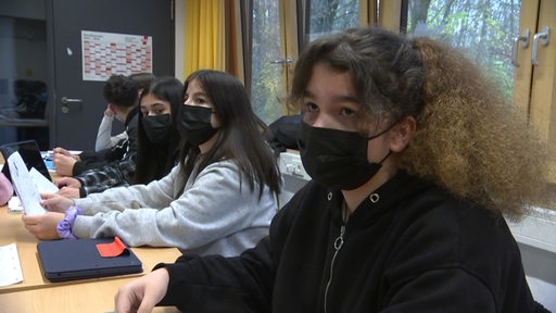 Schülerinnen und Schüler in einem Klassenraum. Drei SChülerinnen gucken in die Kamera.