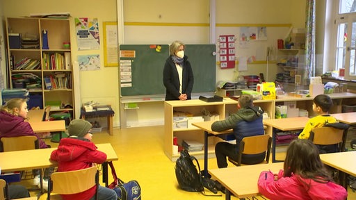 Ein Klassenzimmer mit Schülern, die mit Abstand zueinander sitzen und die Lehrerin, die eine Mund-Nasen-Bedeckung trägt.