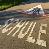 Ein dreieckiges Schild mit Kindern und Schriftzug "Schule" auf einer Straße vor einer Dorfschule.