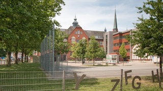 Hinter einem Zaun sind ein Schulhof und Gebäude zu sehen.