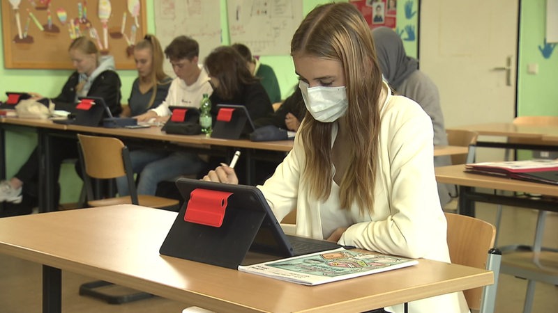 Eine Schülerin sitzt mit Maske in einem Klassenraum vor ihrem Tablet. Im Hintergrund sitzen noch mehr Schüler.