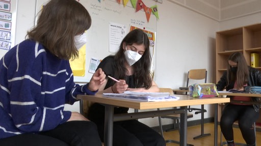 Zwei Schülerinnen in der Schule beim Lernen, die Lehrerin hilft ihnen. Sie tragen alleine eine Maske.