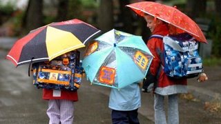 Schulkinder gehen mit Schulranzen und Regenschirm durch den Regen