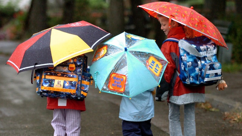 Schulkinder gehen mit Schulranzen und Regenschirm durch den Regen