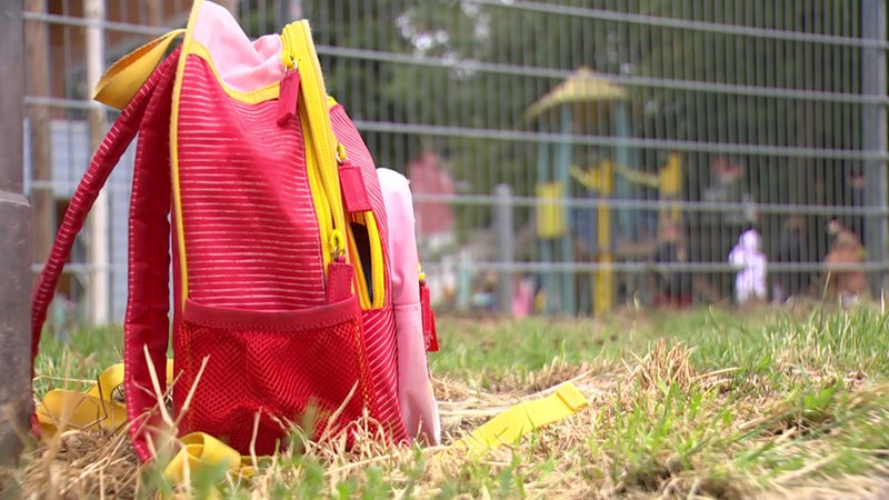 Ein Schulranzen steht vor dem Zaun eines Kita-Spielplatzes.