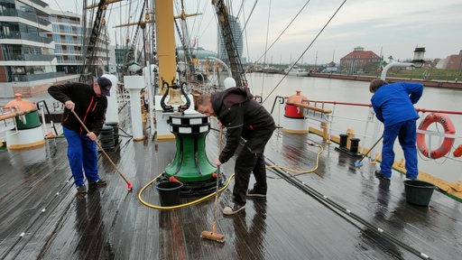 Drei Männer schrubben das Deck eines Schiffes.