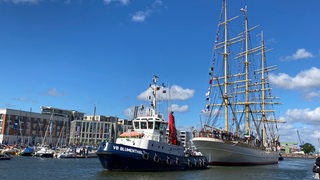 Ein Schlepper zieht ein Segelschiff in einen Hafen.