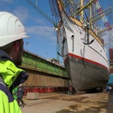 Ein Arbeiter mit Helm und gelber Jacke schaut auf die "Schulschiff Deutschland", ein weißes Segelschiff.