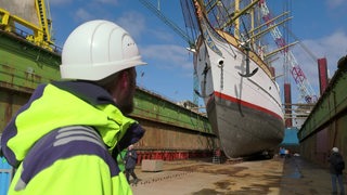 Ein Arbeiter mit Helm und gelber Jacke schaut auf die "Schulschiff Deutschland", ein weißes Segelschiff.