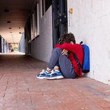 Ein Grundschüler sitze mit seinem Rucksack, den Kopf zwischen den Armen, vor der Schule.