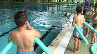 Ein paar Kinder stehen mit Poolnudeln am Beckenrand in einem Schwimmbad.