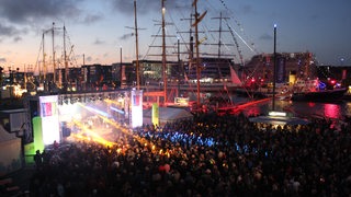 Eine beleuchtete Bühne am Hafenbecken mit Schiffen und Publikum in der Dämmerung