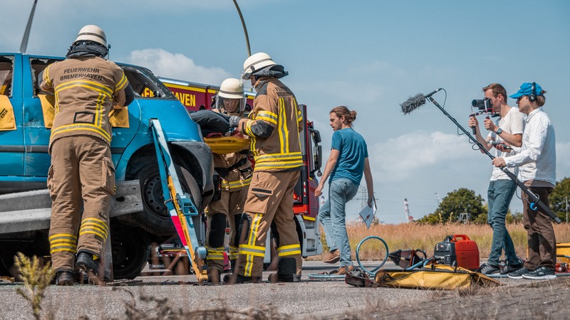 Ein Kamera-Team filmt Feuerwehrleute beim Einsatz an einem Auto.