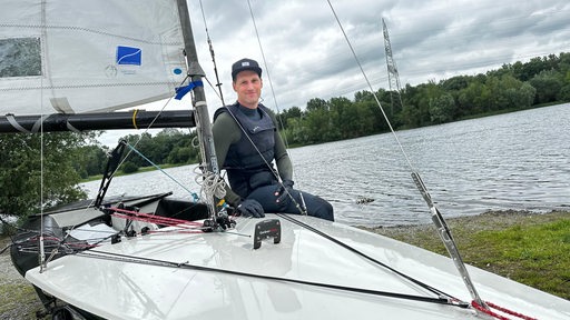 Der Segler Christoph Hohmeier posiert lächelnd an Land vor seinem Boot der Contender Klasse.