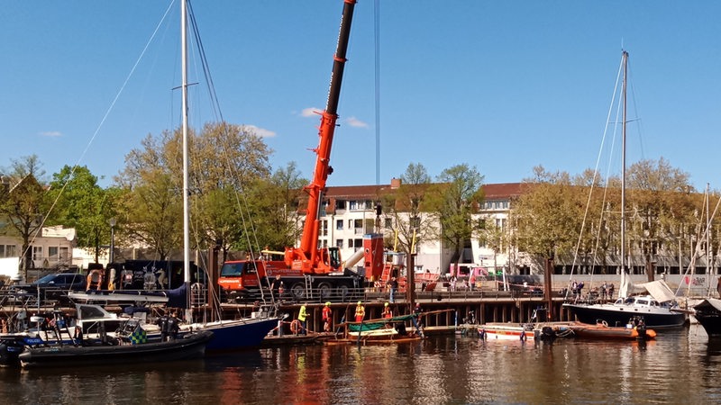Ein Hafen, am Festland steht ein Kran; mehrere Männer auf bergen ein Boot.