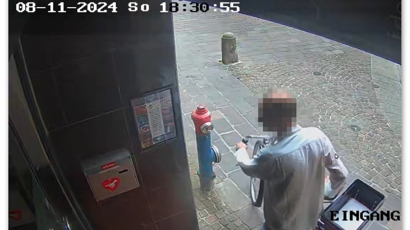 Ein Mann wird von einer Überwachungskamera gefilmt, während er ein Fahrrad schiebt. Kopf ist verpixelt