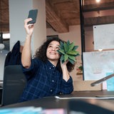 Eine Frau sitzt vor einem Laptop und macht ein Selfie, während sie eine Pflanze in der Hand hält.