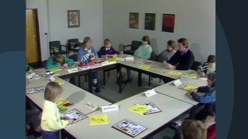 In einem Seminarraum sitzen Kinder und Erwachsene an Tischen. Auf den Tischen liegen Prospekte und Bücher.