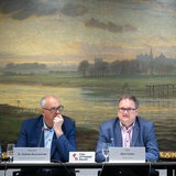 Das Bild zeigt drei Menschen, Bürgermeister Andreas Bovenschulte, Finanzsenator Björn Fecker sowie Wirtschaftsministerin Kristina Vogt, die gemeinsam an einem Tisch mit Mikrofonen vor einem landschaftlichen Gemälde, das Bremen zeigt.