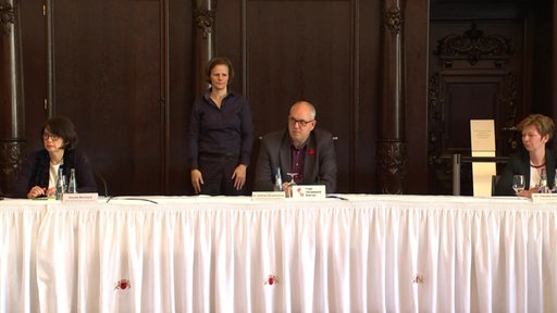 Drei Menschen sitzen vor Mikrofonen an einem Tisch