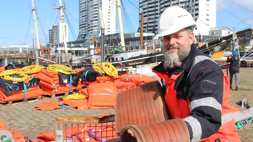 Ein Mann mit Arbeitskleidung und Helm steht vor dem Segelschiff und zeigt einen breiten Gurt.