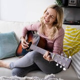 Eine Frau sitzt zuhause auf dem Sofa, spielt Gitarre und singt dazu.