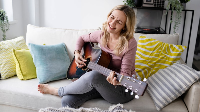 Eine Frau sitzt zuhause auf dem Sofa, spielt Gitarre und singt dazu.