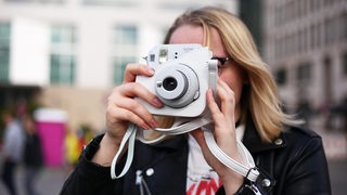 Eine junge Frau fotografiert vor Gebäuden mit einer Sofortbildkamera.