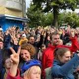 Zahlreiche Menschen haben sich auf einer Straße versammelt und heben eine Hand
