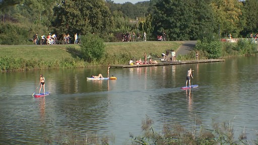 An einem Sommertag sind viele Menschen am Werdersee. Einige von ihnen sind auf dem Steg oder auf dem Wasser beim Stand-Up paddeln. 