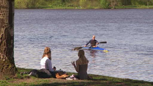 Drei Mädchen sitzen an dem Werder-See Ufer und im Hintergrund rudert ein Mann auf dem Gewässer.
