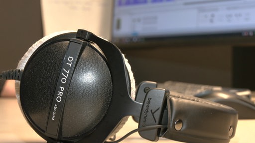 Große Kopfhörer liegen auf einem Schreibtisch. Im Hintergrund ist ein Computer zu sehen.