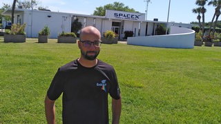 Ein Mann im T-Shirt steht vor dem Firmengebäude von Spacex, Umgebung zeigt Palmen
