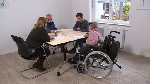 Es sitzen vier Personen an einem Tisch, darunter eine in einem Rollstuhl.