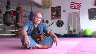 Klaus Handke sitzt im Schneidersitzt auf dem Boden in einem Kampfsportstudio.