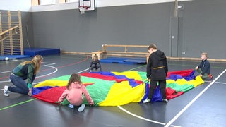 Erstklässler umringen in einer Turnhalle ein buntes rundes Tuch.