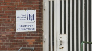 Ein Schild mit der Aufschrift Bibliothek im Strafvollzug an einer Backsteinmauer neben einer Tür.