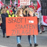 Mit Schildern und Transparenten demonstrieren Teilnehmende während eines Warnstreiks. Auf einem Schildern steht die Aufschrift „Stadtstaatenzulage jetzt“.