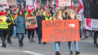 Mit Schildern und Transparenten demonstrieren Teilnehmende während eines Warnstreiks. Auf einem Schildern steht die Aufschrift „Stadtstaatenzulage jetzt“.