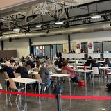 Zahlreiche Menschen in einem Saal sitzen an Tischen.