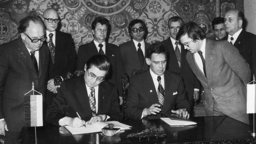 Zehn Männer stehen an einem Tisch vor einem Wandteppich. Zwei von ihnen sitzen an einem Tisch und unterzeichnen einen Vertrag.