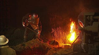 Ein Stahlarbeiter in silberner Ausrüstung schaufelt Schlacke. Rechts ein Feuer.