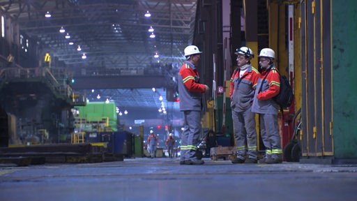 Drei Mitarbeiter von ArcelorMittal stehen im Stahlwerk. 