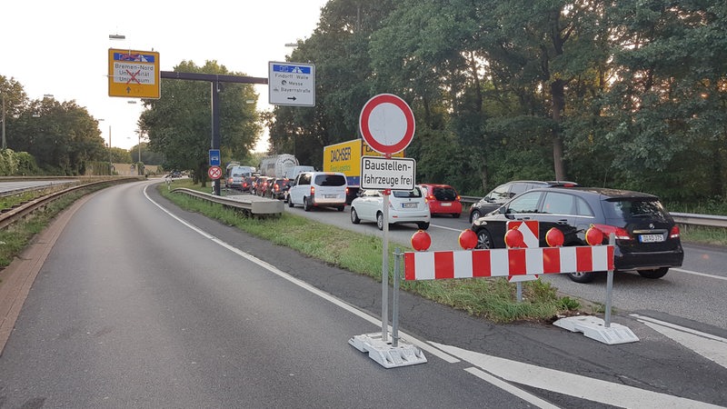 Ein Baustellenschild steht auf einer Straße, auf der danebenliegenden Spur, stehen Autos im Stau.
