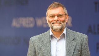 Ein Mann im Sakko steht vor einer blauen Wand und blickt in die Kamera.