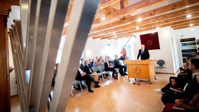 Ein Mann im Anzug steht hinter einem hölzernen Pult mit Davidstern und spricht zu sitzenden Zuschauern.